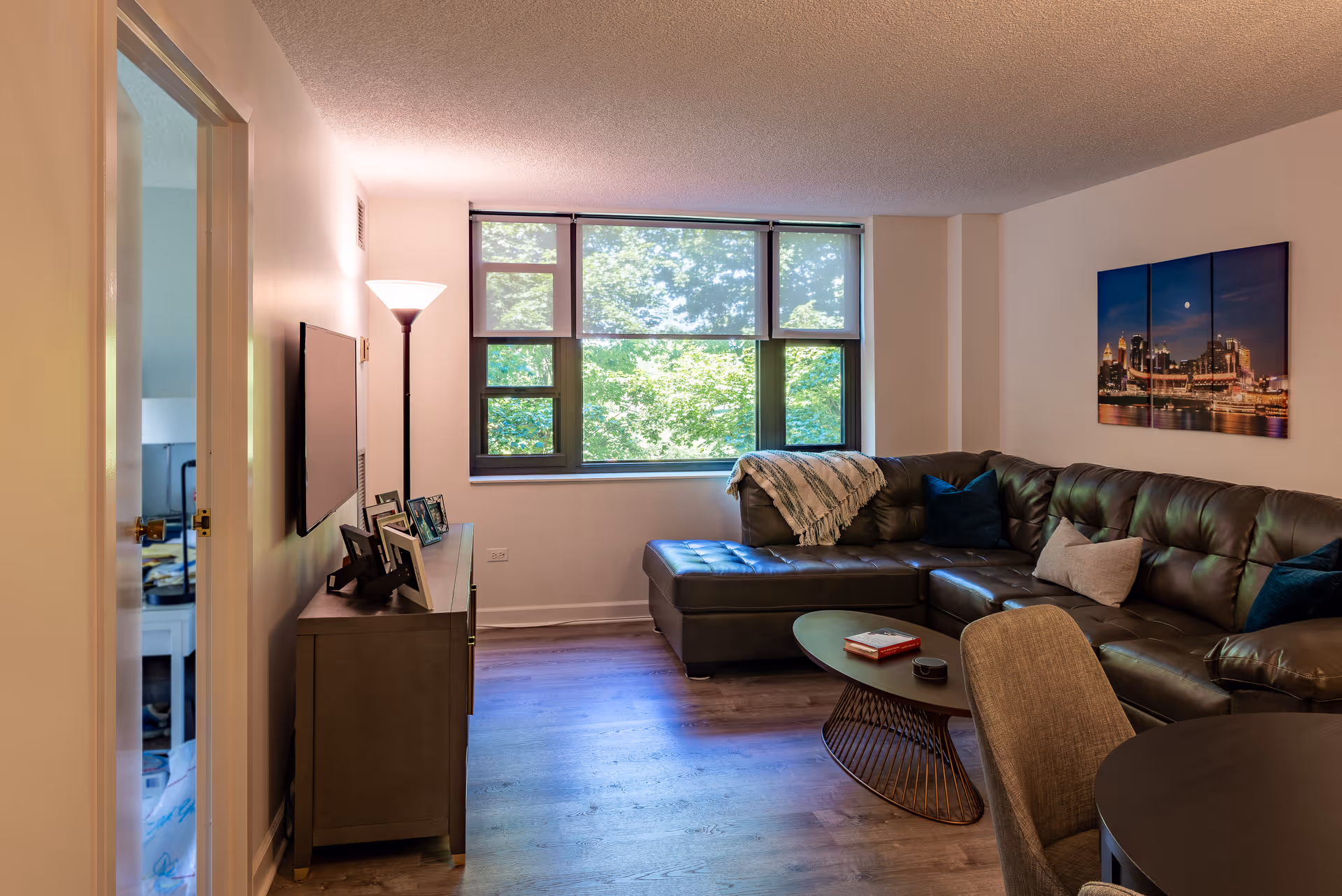 A cozy living room with a large dark brown leather sectional sofa adorned with pillows and a throw blanket. A modern oval coffee table sits in front of the sofa with a book and a small item on it. A floor lamp stands next to a TV mounted on the wall above a cabinet with framed photos. Large windows let in natural light and show green trees outside. A piece of wall art depicting a city skyline at night hangs above the sofa. The room has wooden flooring and neutral-colored walls.