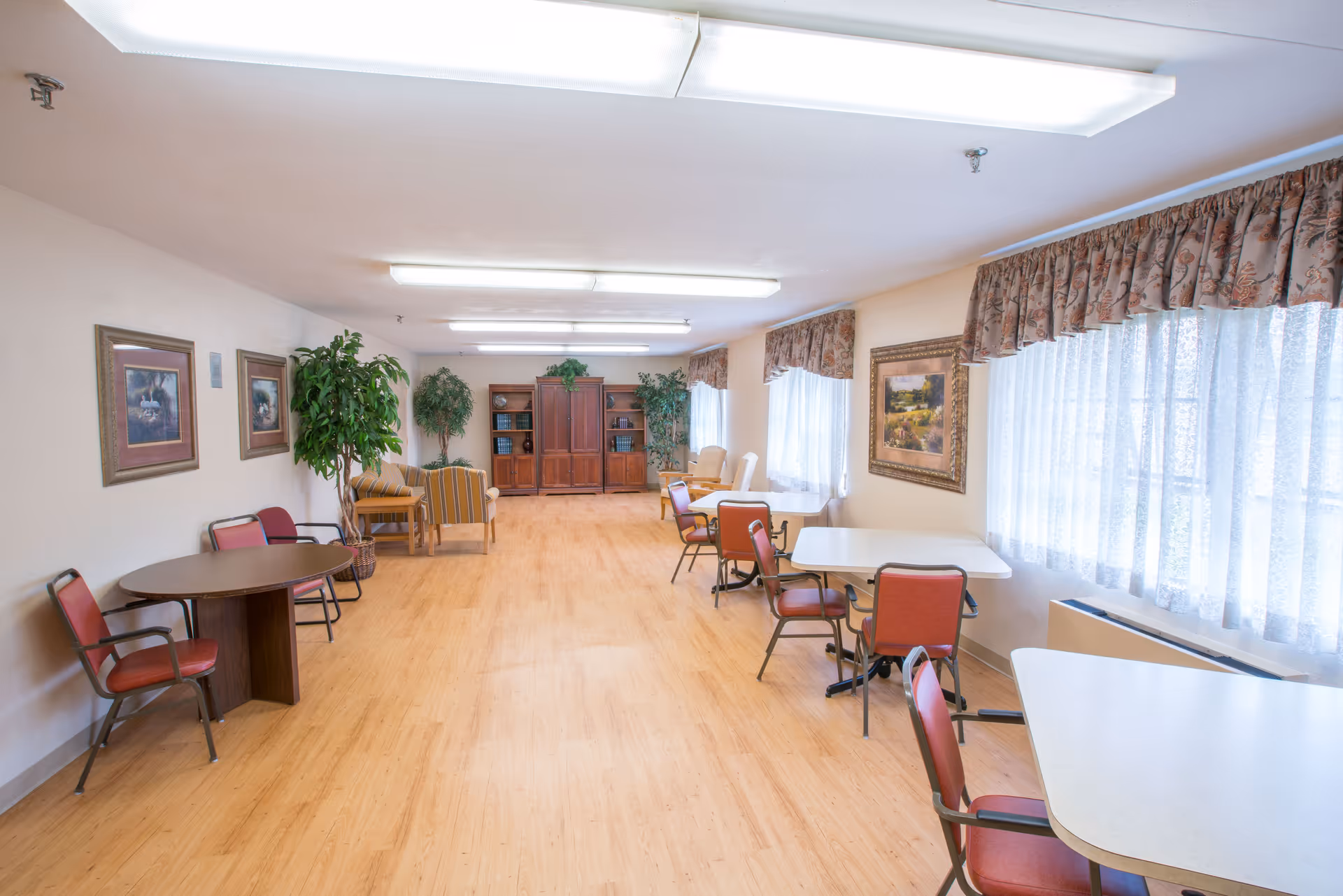 A bright and spacious common area with multiple tables and chairs arranged along the walls. The room has large windows with sheer curtains and floral valances, framed artwork on the walls, potted plants, and a wooden cabinet with shelves at the far end. The floor is light wood, and the ceiling has fluorescent lighting.