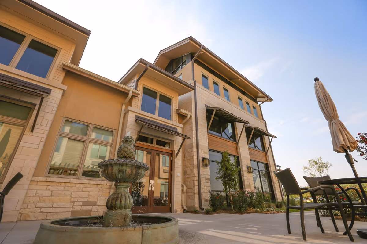 Exterior view of a multi-story building with large windows and a stone facade under a clear sky. In the foreground, there is a decorative water fountain and outdoor seating with a closed umbrella.