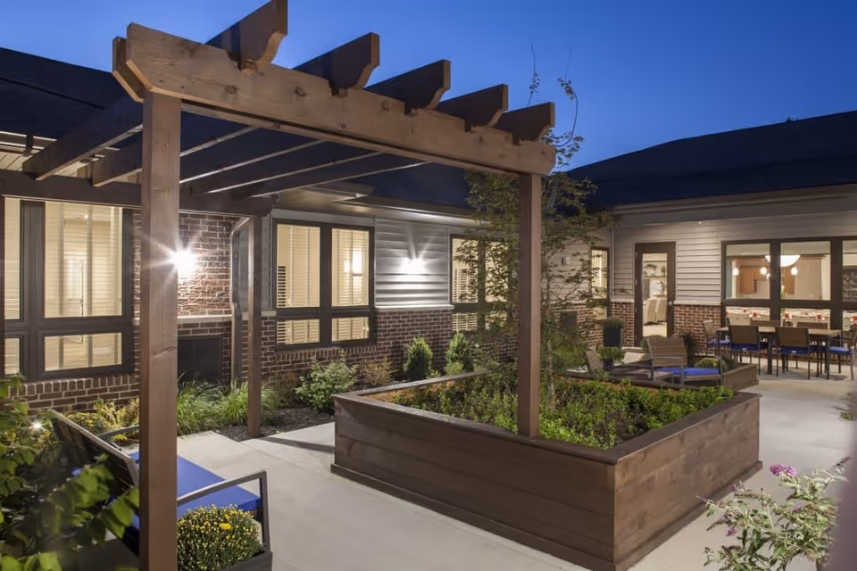 Outdoor courtyard area at dusk with a wooden pergola, raised garden beds, patio seating, and a building with large windows and exterior lighting.
