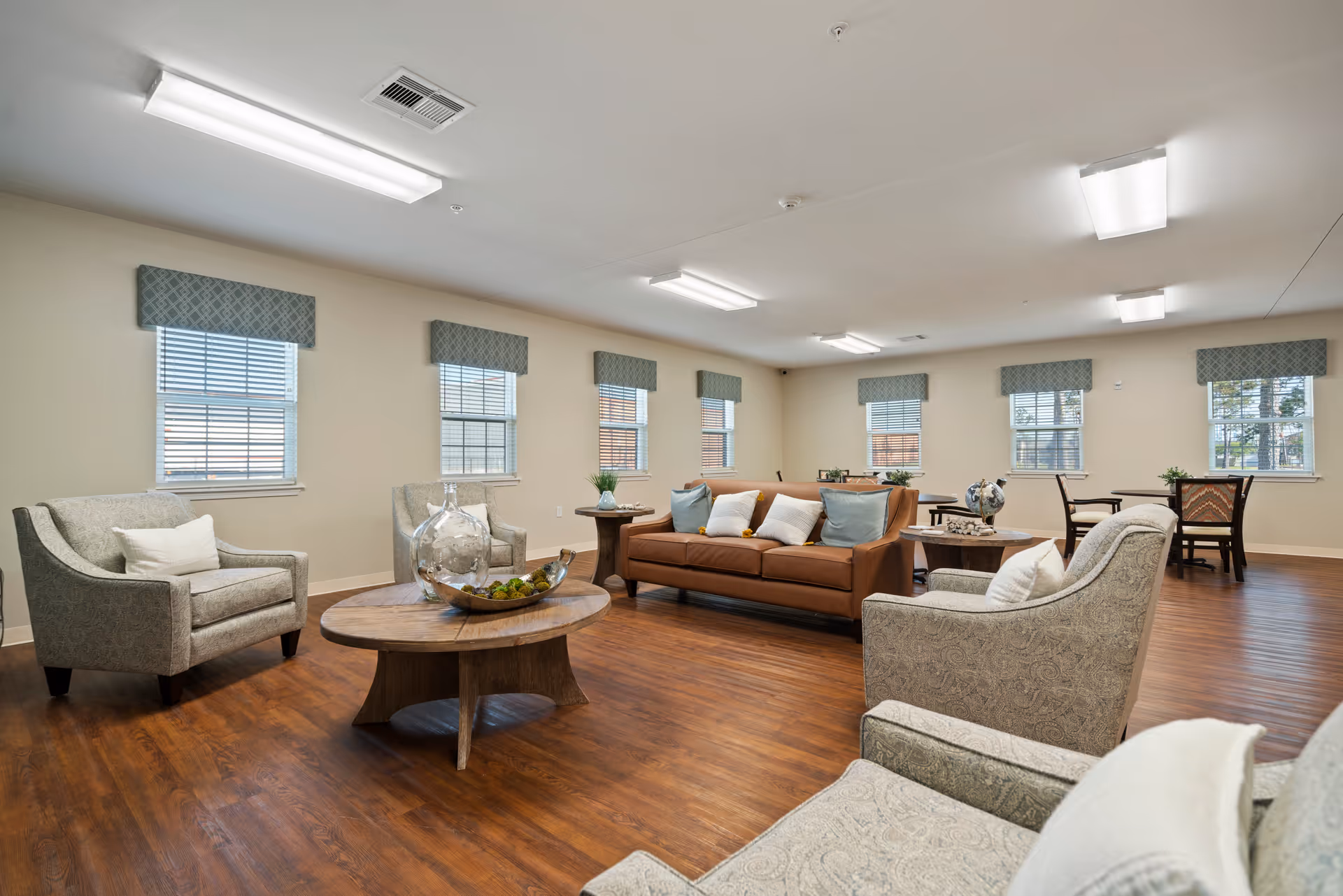 A spacious living room with wooden flooring, featuring a brown leather sofa with multiple cushions, several patterned armchairs, round wooden coffee tables with decorative items, and multiple windows with blue patterned valances allowing natural light to fill the room.