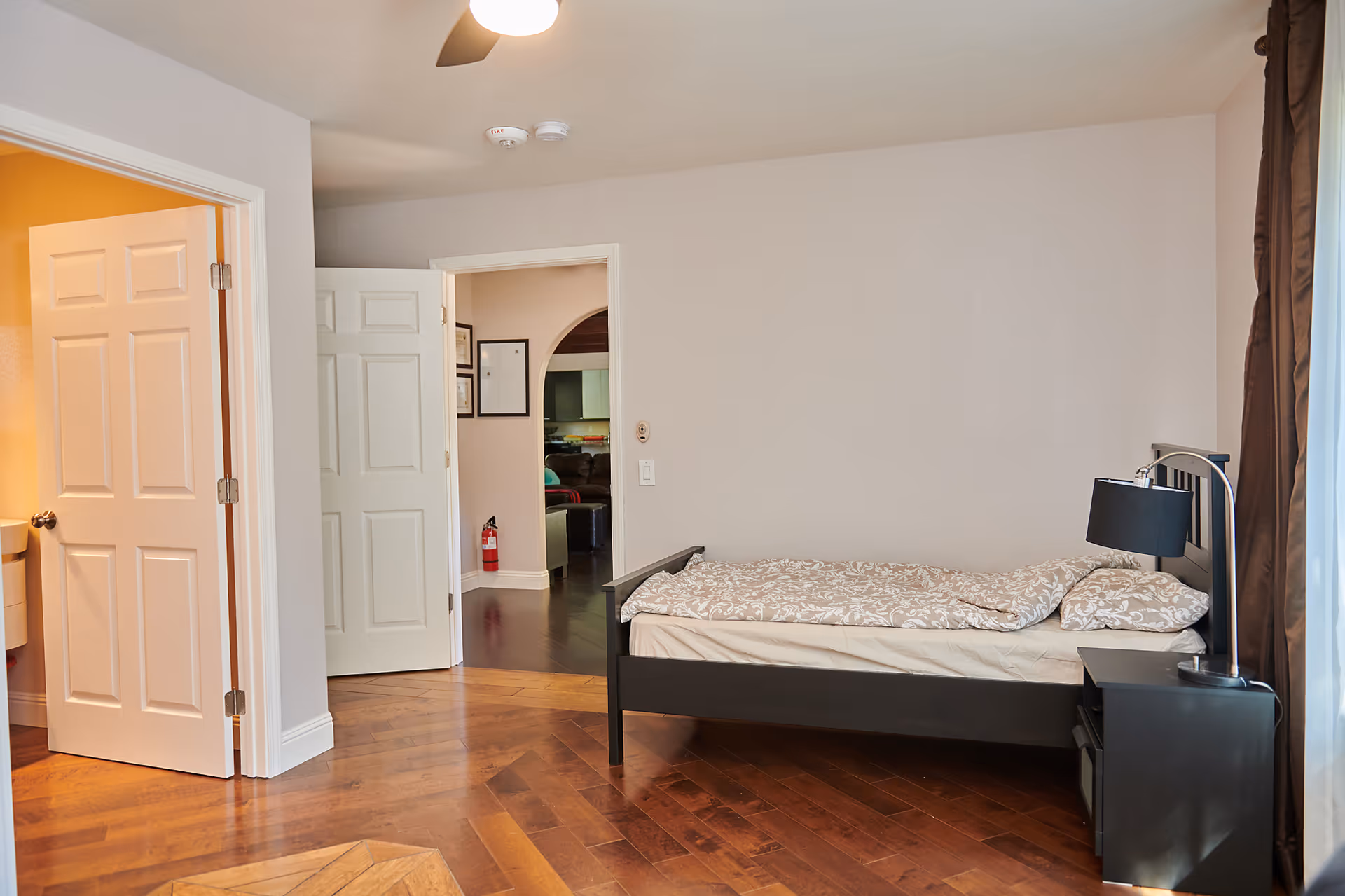 A bedroom with a single bed against the right wall, covered with a patterned beige and white comforter and pillow. Next to the bed is a black nightstand with a black lamp. The room has wooden flooring and light-colored walls. There is an open door on the left leading to a bathroom with a sink visible, and another open door in the center leading to a hallway with dark flooring and a fire extinguisher mounted on the wall.