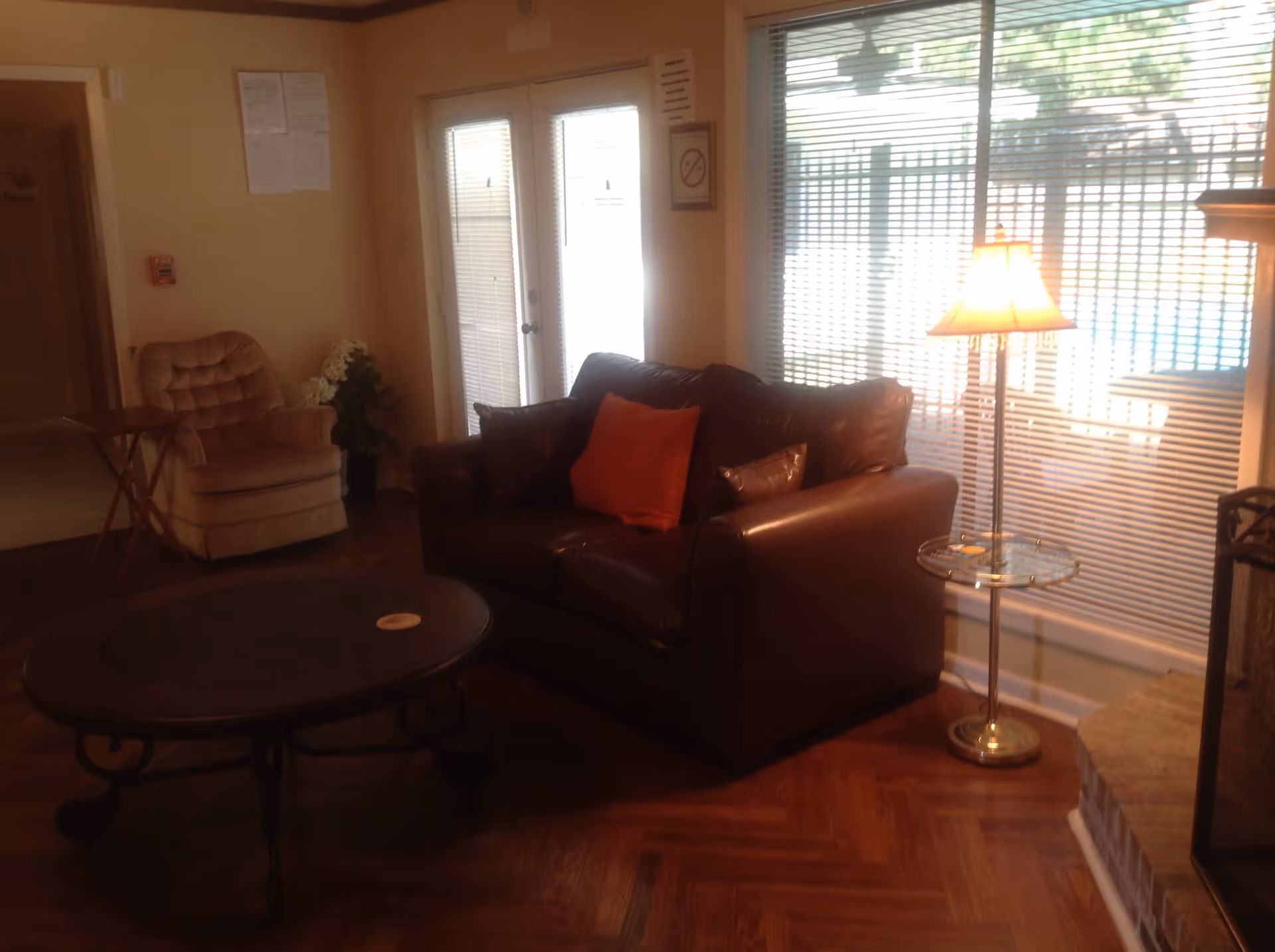 A cozy living room with a dark brown leather loveseat featuring a red throw pillow, a round coffee table with ornate legs, a beige armchair, a small wooden side table, a floor lamp with a glass tray, and large windows with blinds letting in natural light.