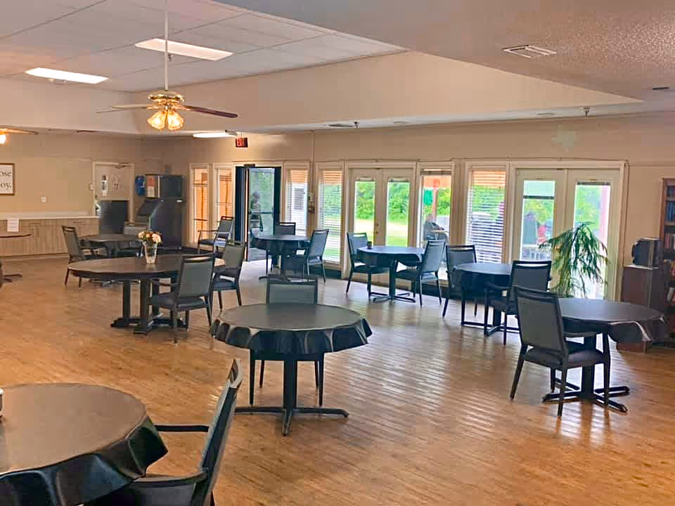 A spacious dining room with multiple round tables covered with black tablecloths and surrounded by chairs. The room has wooden flooring, large windows and glass doors letting in natural light, a ceiling fan with lights, and a few decorative plants. The walls are beige, and there is a water dispenser and some wall decor visible in the background.