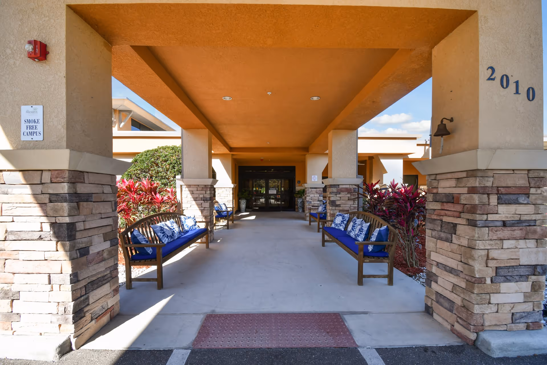 Covered entrance area of The Preserve at Dunedin with stone pillars and wooden benches with blue cushions and patterned pillows on either side. There are red-leafed plants along the sides and double glass doors at the end of the walkway under a tan ceiling.