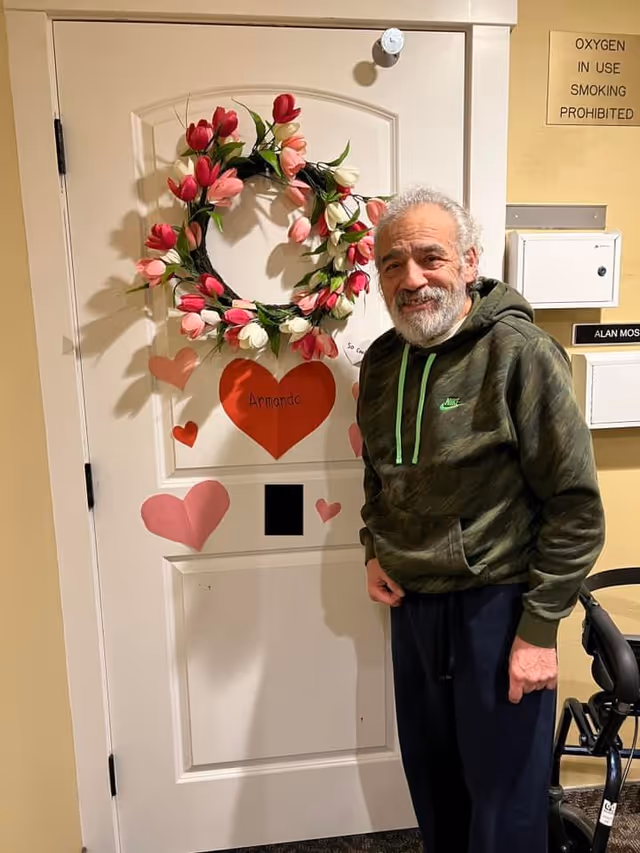 An elderly man with gray hair and a beard stands smiling next to a white door decorated with a wreath of pink and white flowers and several paper hearts, one of which has the name 'Armando' written on it. The man is wearing a green camouflage hoodie and dark pants. A walker is partially visible to the right. On the wall behind him, there is a sign that reads 'OXYGEN IN USE SMOKING PROHIBITED' and a nameplate with 'ALAN MOS' visible.