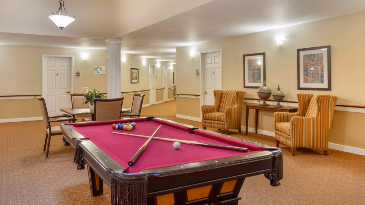 Interior view of a senior living facility common area featuring a red pool table with two pool cues and balls arranged on it. In the background, there are two striped armchairs with a wooden table between them, decorated with two ornamental vases. A small dining table with four chairs and a plant centerpiece is also visible. The walls are beige with framed artwork, and the hallway has several doors and wall-mounted lights.