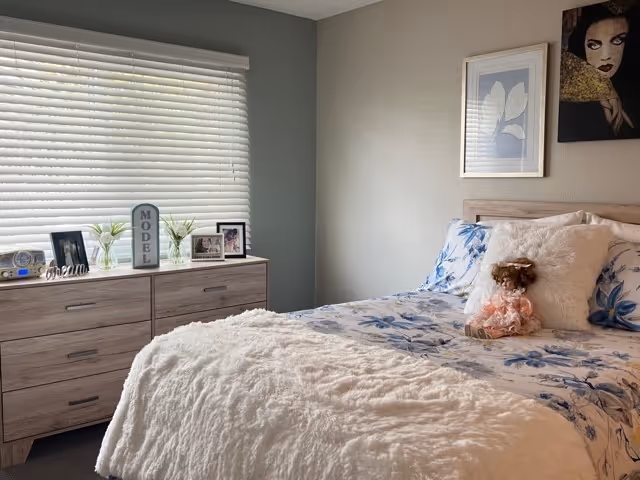 A cozy bedroom with a bed covered in a white fluffy blanket and floral sheets. A doll is placed on the bed. Next to the bed is a wooden dresser with framed photos, small vases with flowers, and a decorative sign that reads 'MODEL'. The room has a window with closed blinds and two framed artworks on the wall above the bed.