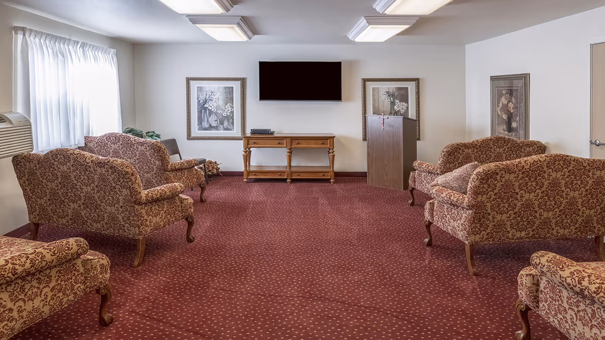 Carpeted common living room with patterned sofas arranged facing a wall-mounted TV and a small wooden table.