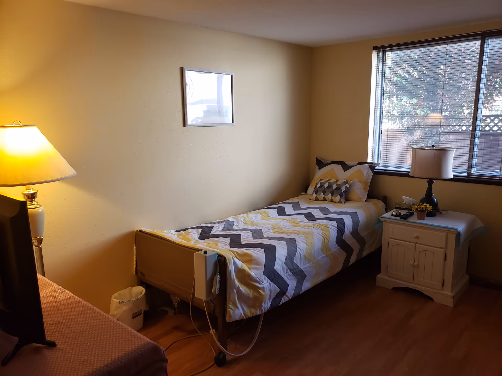 A small bedroom with a single bed covered in a yellow, gray, and white chevron-patterned blanket and matching pillows. Next to the bed is a white nightstand with a lamp, a small potted plant, a tissue box, and a remote control. There is a large window with blinds behind the nightstand, and a lamp on a table is visible on the left side of the image. The room has wooden flooring and light-colored walls.