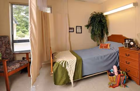 A bedroom in a senior care facility with a bed, dresser, curtains, and a leafy plant.