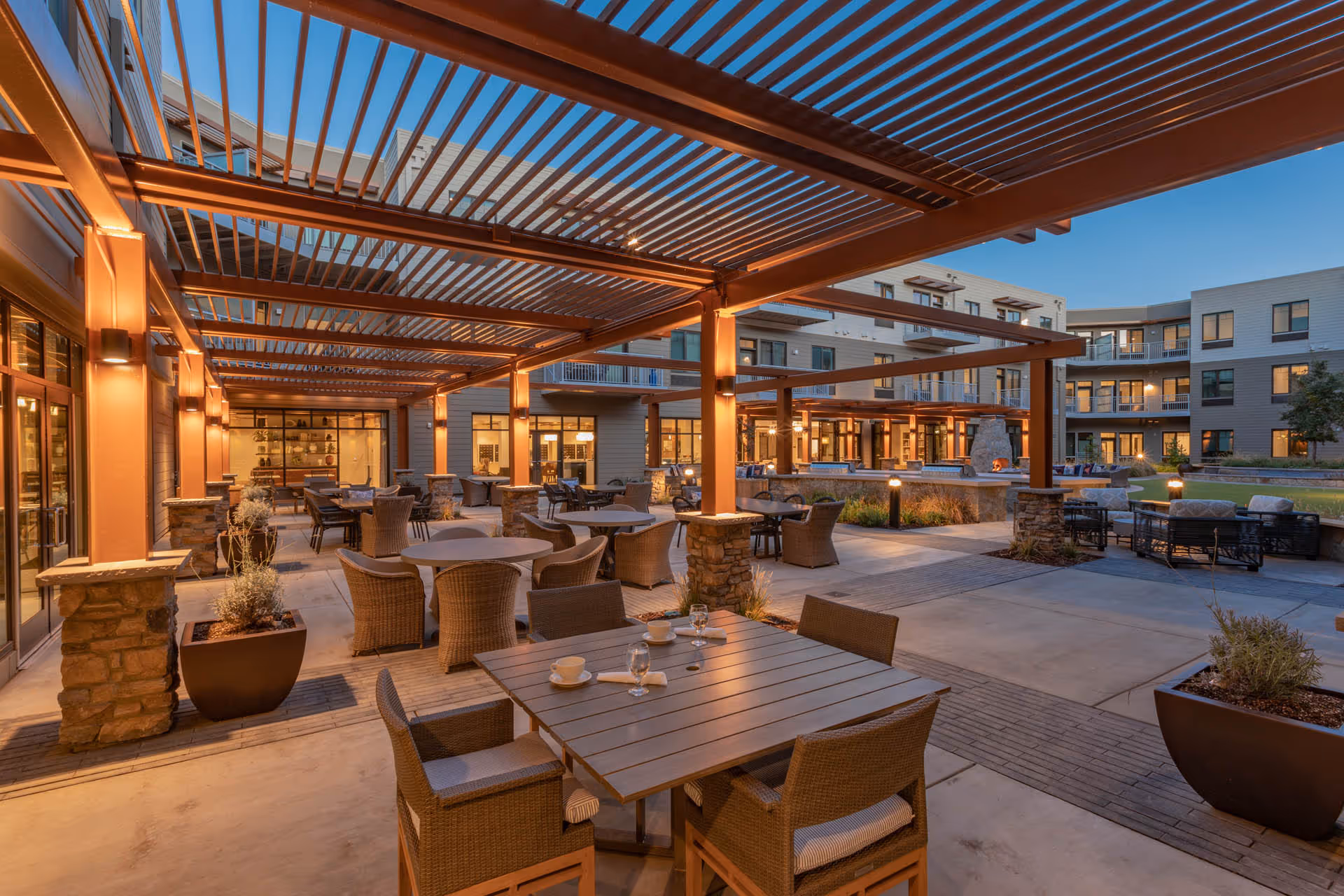 Outdoor patio area at The Watermark at Napa Valley with multiple tables and wicker chairs under a pergola structure, surrounded by a modern building with large windows and balconies, illuminated by warm lighting at dusk.