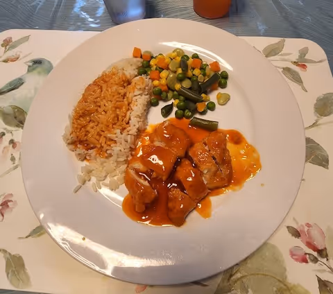 A white plate with a serving of rice mixed with tomato sauce, a portion of mixed vegetables including peas, corn, carrots, and green beans, and pieces of chicken covered in a reddish-orange sauce. The plate is on a floral placemat with a glass of water and a glass of orange drink in the background.