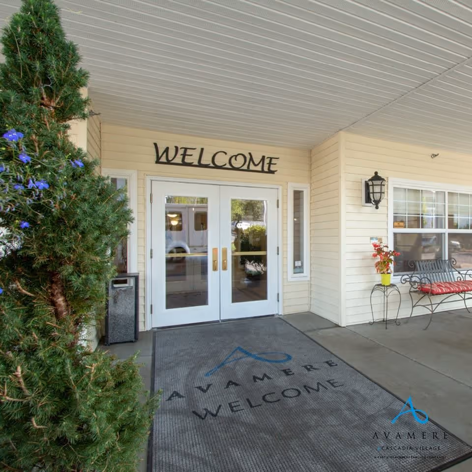 Entrance to Cascadia Village Senior Living by Cogir with double glass doors under a covered porch. Above the doors is a sign that says 'WELCOME'. There is a large welcome mat on the ground with the Avamere logo and the word 'WELCOME'. To the left is a green bush with purple flowers, and to the right is a bench with a red cushion and a small table holding a potted plant.