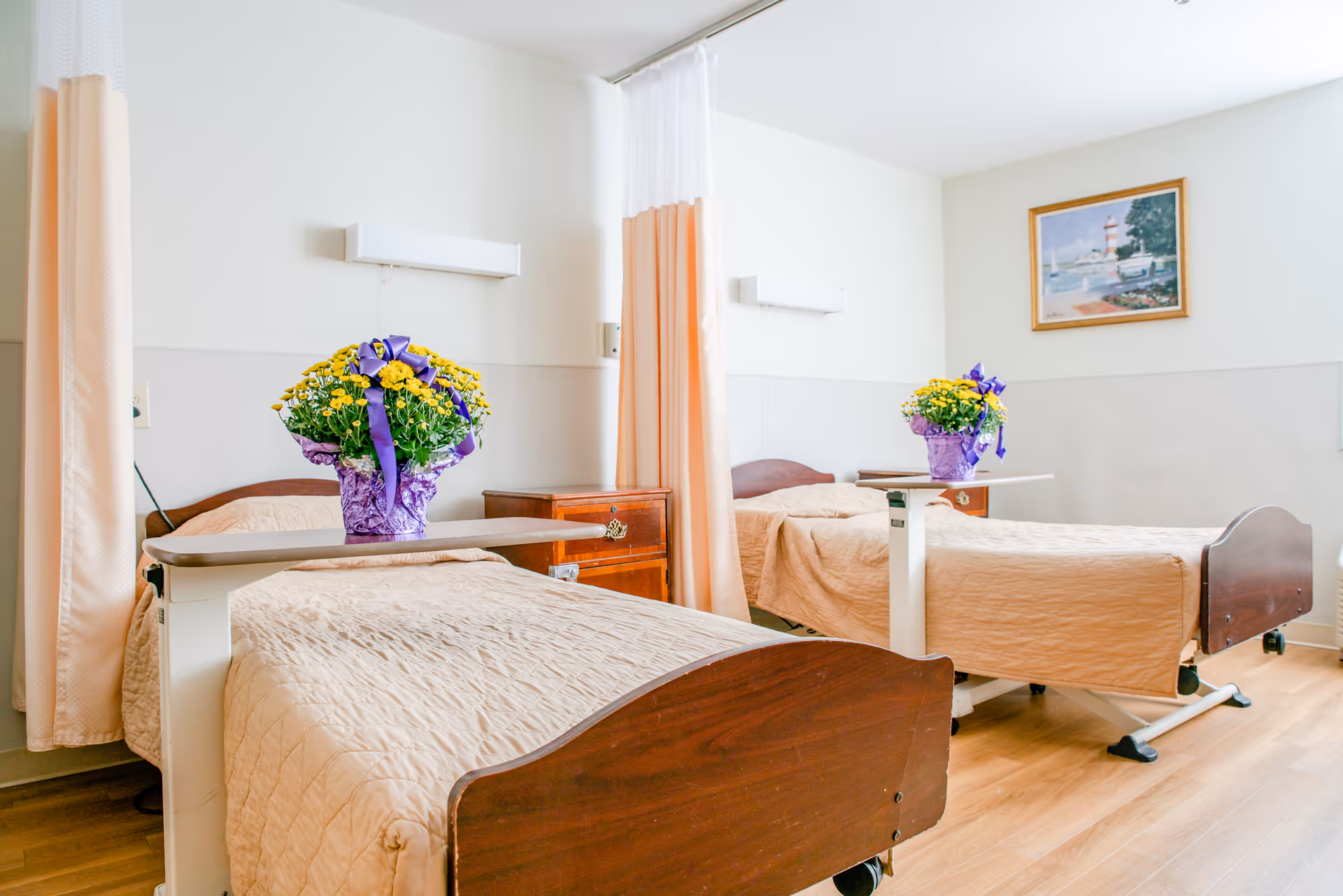 A bright room with two hospital-style beds separated by a privacy curtain. Each bed has a beige quilted blanket and a small overbed table holding a purple pot with yellow flowers and a purple ribbon. There is a wooden nightstand between the beds and a framed painting of a lighthouse on the wall.