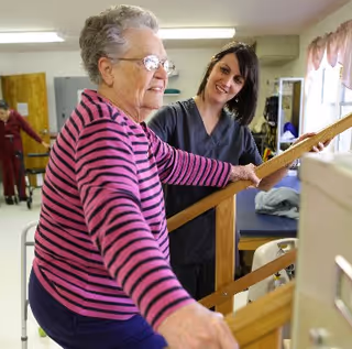 An elderly woman in a pink and black striped shirt is holding onto a wooden handrail while a female caregiver in gray scrubs supports and encourages her in a bright room with exercise equipment.