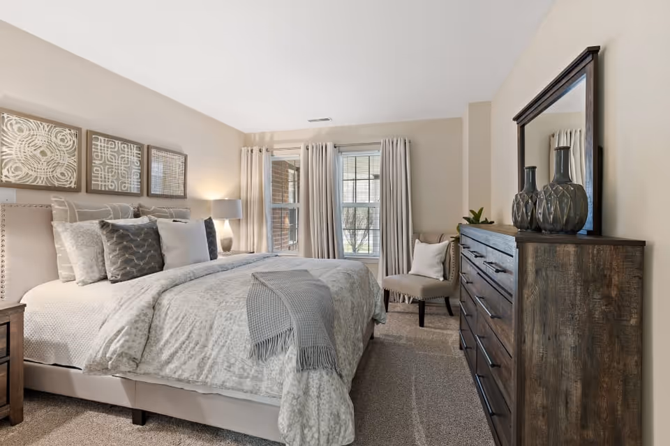 A cozy bedroom with a large bed covered in light gray and white bedding, multiple decorative pillows, and a gray throw blanket. On the left side of the bed is a wooden nightstand with a lamp. Above the bed are three framed abstract art pieces. Two windows with beige curtains allow natural light into the room. To the right, there is a wooden dresser with a large mirror and decorative vases on top. A beige upholstered chair with a white pillow is placed near the windows.
