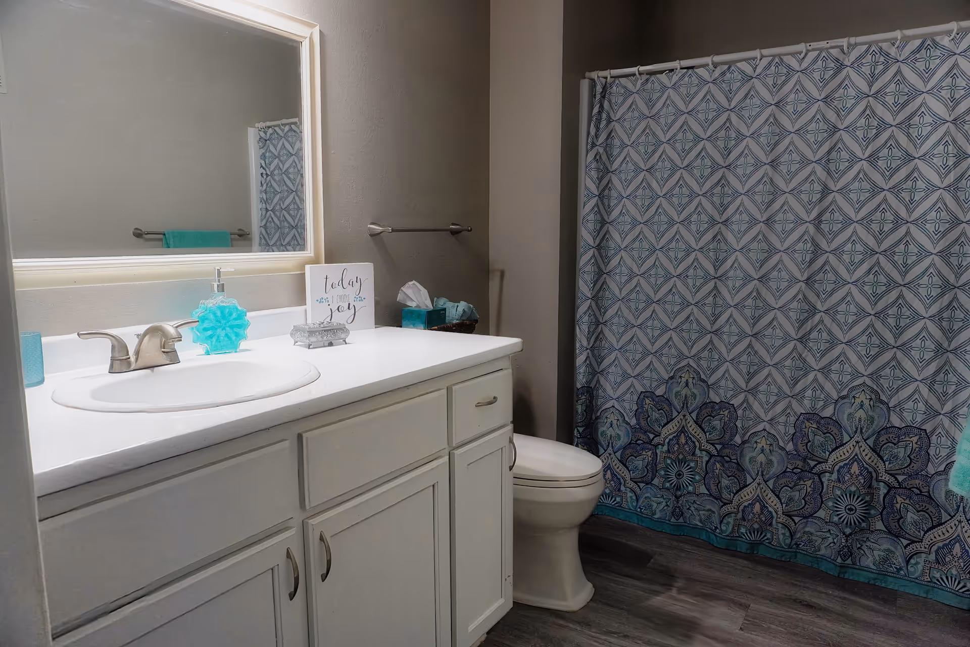A bathroom with a white vanity cabinet, a sink with a silver faucet, a large mirror above the sink, a decorative soap dispenser, a small sign that reads 'today is a good day for joy', a tissue box, a toilet, and a shower with a blue and white patterned curtain.