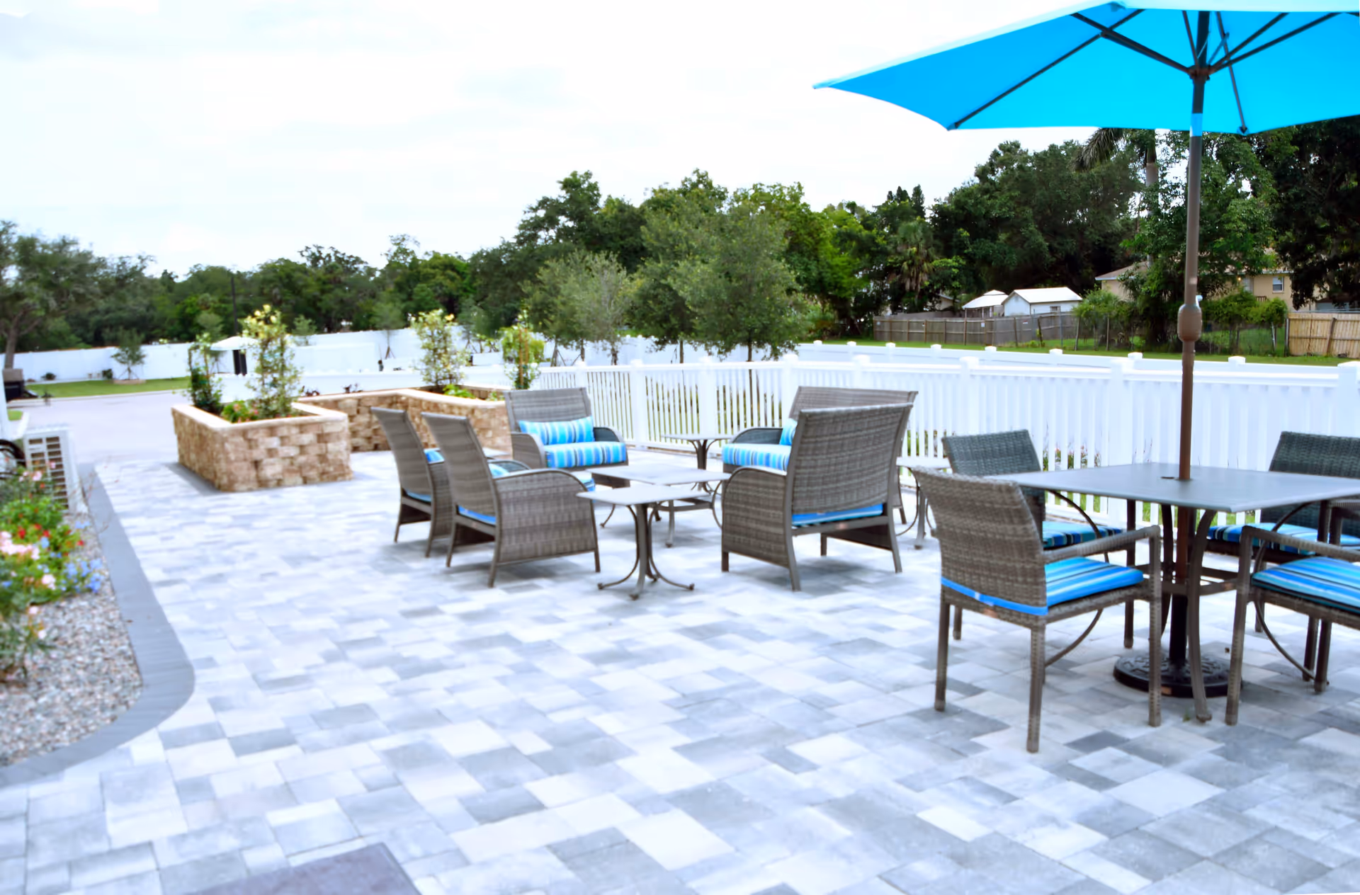 Outdoor patio area with gray and blue cushioned wicker chairs and tables, including one table with a large blue umbrella. The patio is paved with light gray tiles and is surrounded by a white fence with greenery and trees in the background.