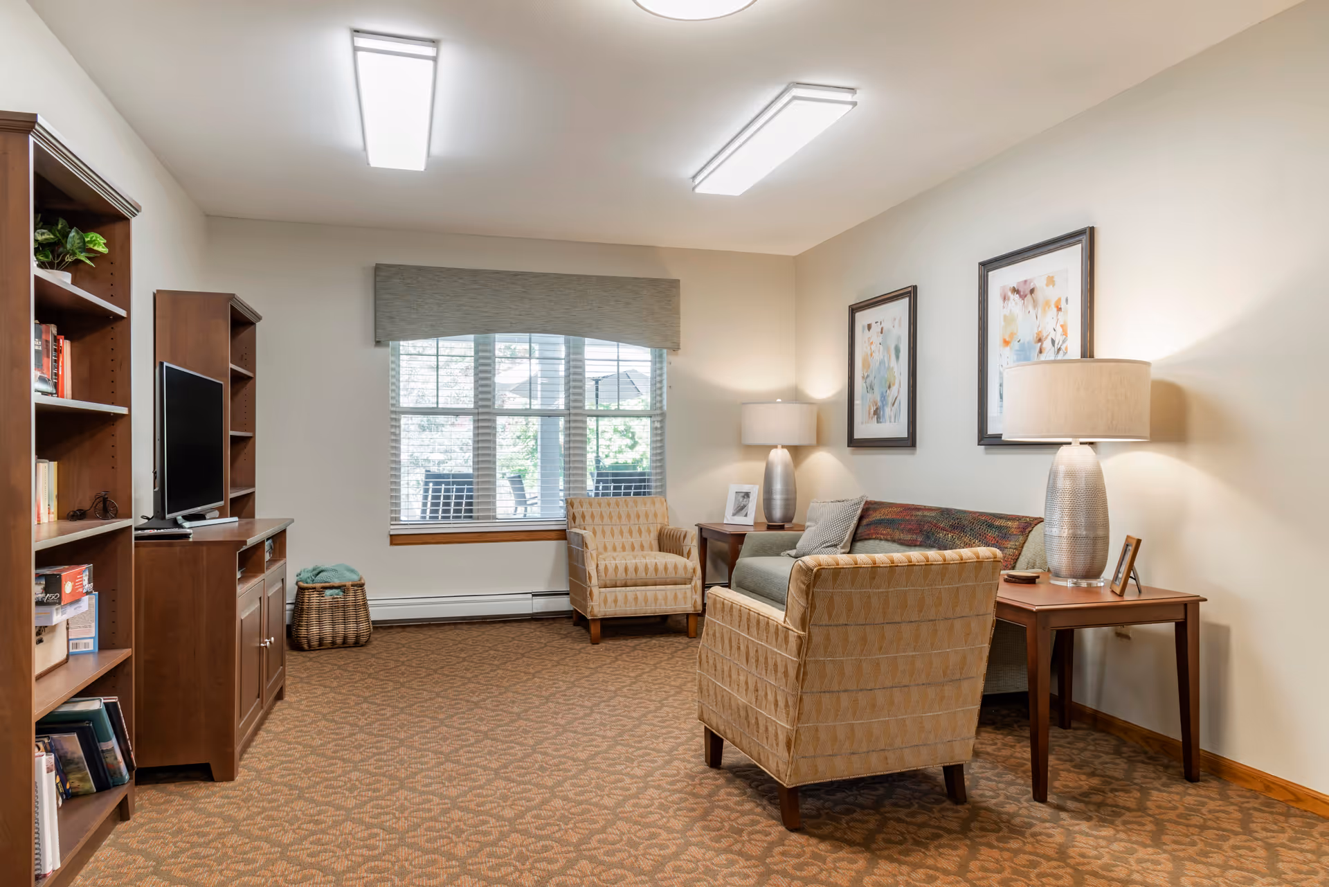 A cozy living room with patterned carpet, two beige armchairs, a gray sofa with a colorful throw, two wooden side tables each with a lamp, framed artwork on the wall, a large window with blinds and a valance, and a wooden entertainment center with a TV and bookshelves.