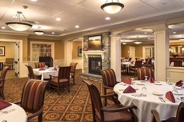 Elegant dining room with round tables set with white linens and burgundy napkins, upholstered chairs, chandeliers, and a central stone fireplace.
