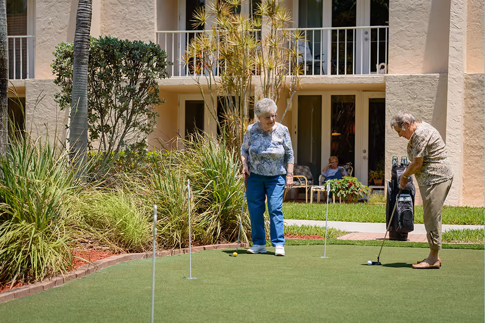 Two elderly women playing on a putting green outside a senior living building while another resident sits on a patio in the background.