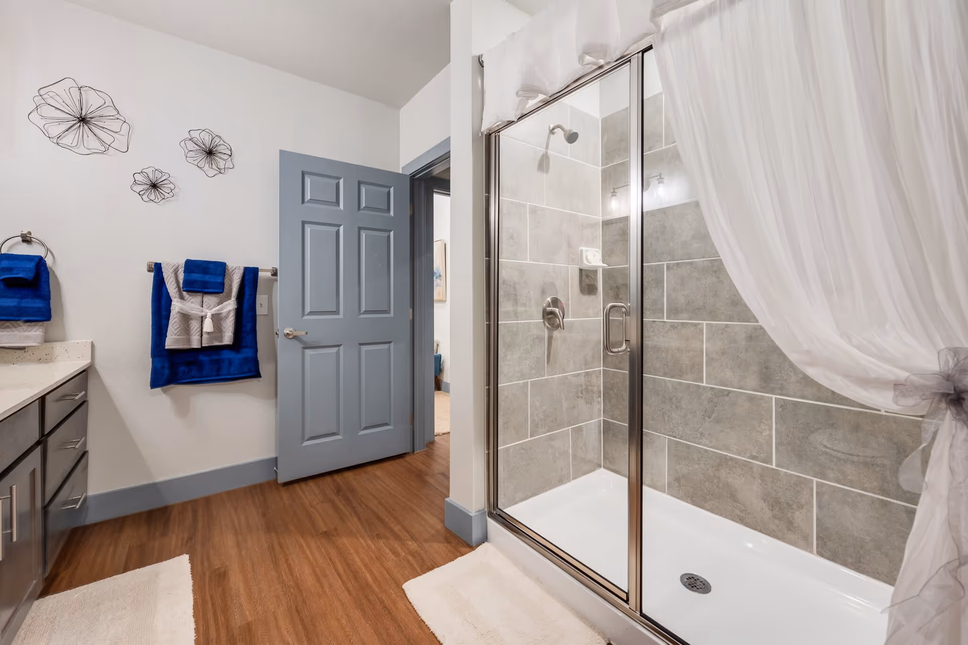 Bright modern bathroom with a glass-enclosed shower, blue door and towels, vanity to the left and wood-look flooring.