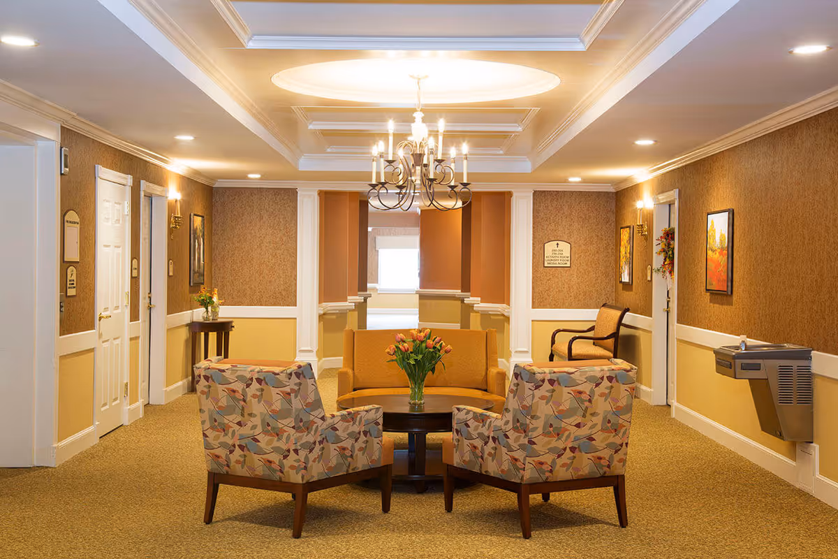 A well-lit sitting area in a senior living facility with a chandelier hanging from a coffered ceiling. The room features a mustard-colored sofa, two patterned armchairs, a round wooden coffee table with a vase of flowers, and additional seating along the walls. The walls are decorated with framed artwork and sconces, and there is a water fountain on the right side.