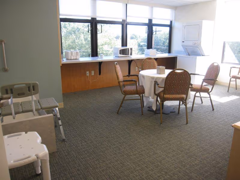 A bright room with large windows, featuring a round table covered with a white tablecloth and surrounded by four patterned chairs. Against the wall is a counter with a microwave, toaster, and folded towels. A stacked washer and dryer unit is in the corner, and two shower chairs are visible on the left side of the image.