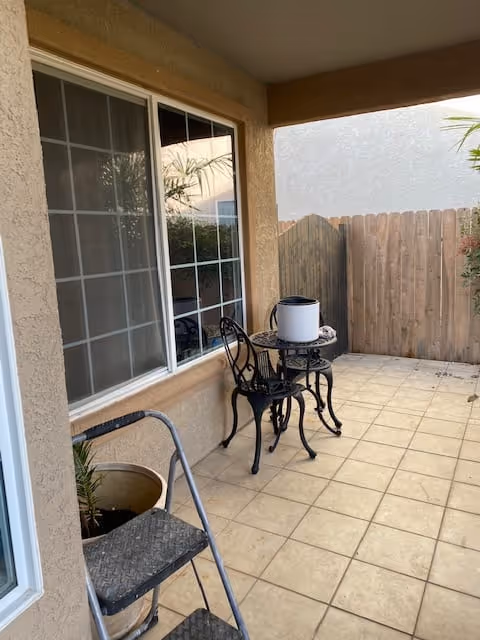 A tiled outdoor patio area with a small black metal table and two matching chairs. There is a white cylindrical object on the table. A wooden fence encloses the patio, and a large window with white framing looks into the building. A metal step ladder and a potted plant are also visible near the window.