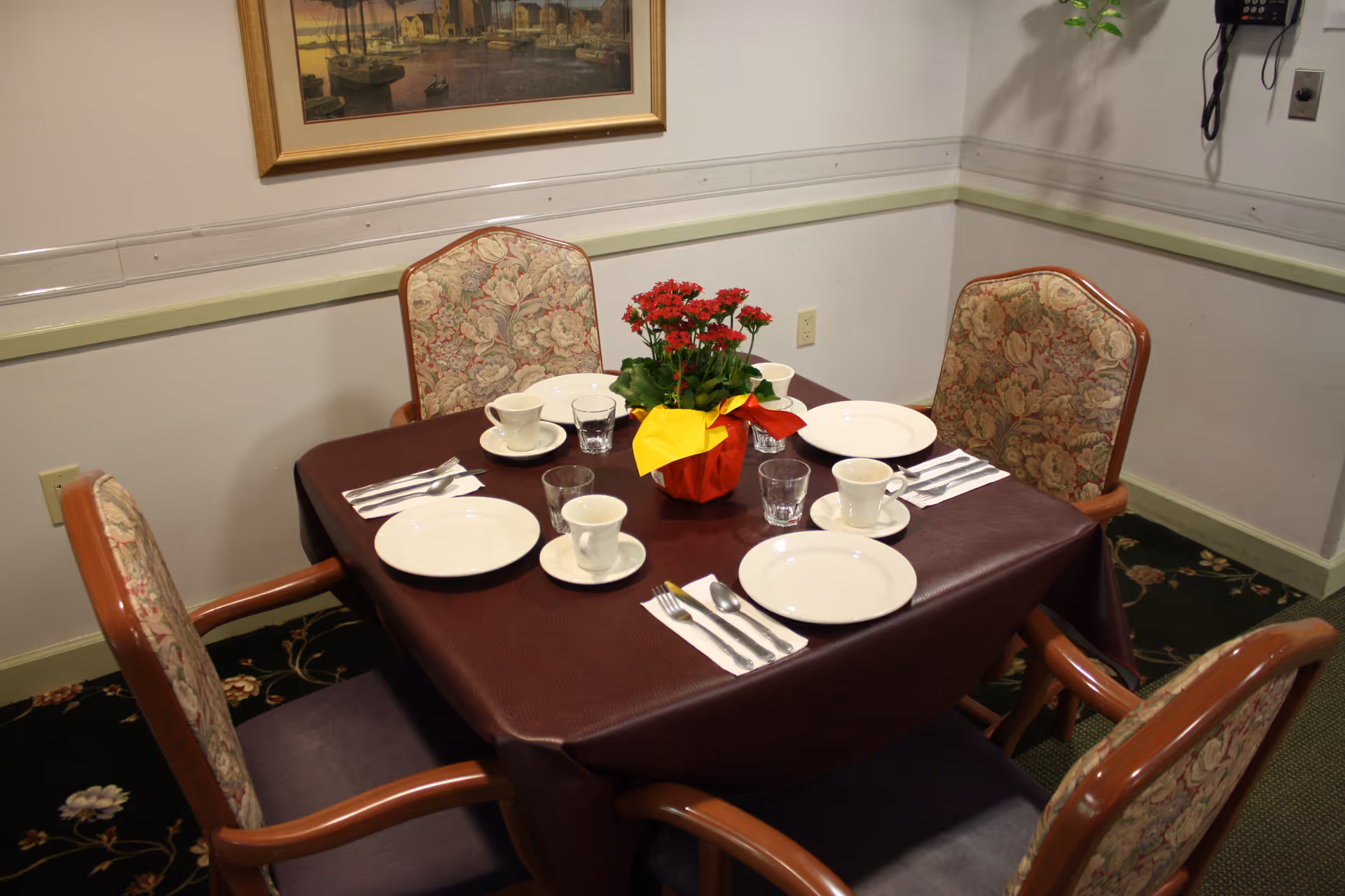 A small square dining table set for four with white plates, cups, utensils and a red potted flower centerpiece in a cozy dining room.