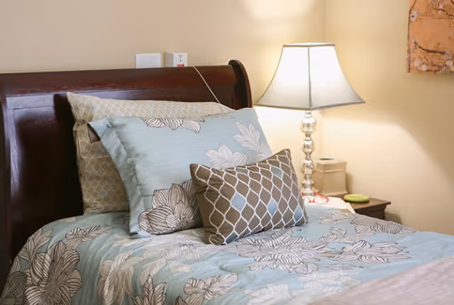 A neatly made bed with a dark wooden headboard, adorned with multiple pillows in various patterns including floral and geometric designs. A bedside table with a decorative lamp and some small items is visible next to the bed. The wall behind is painted a light beige color.
