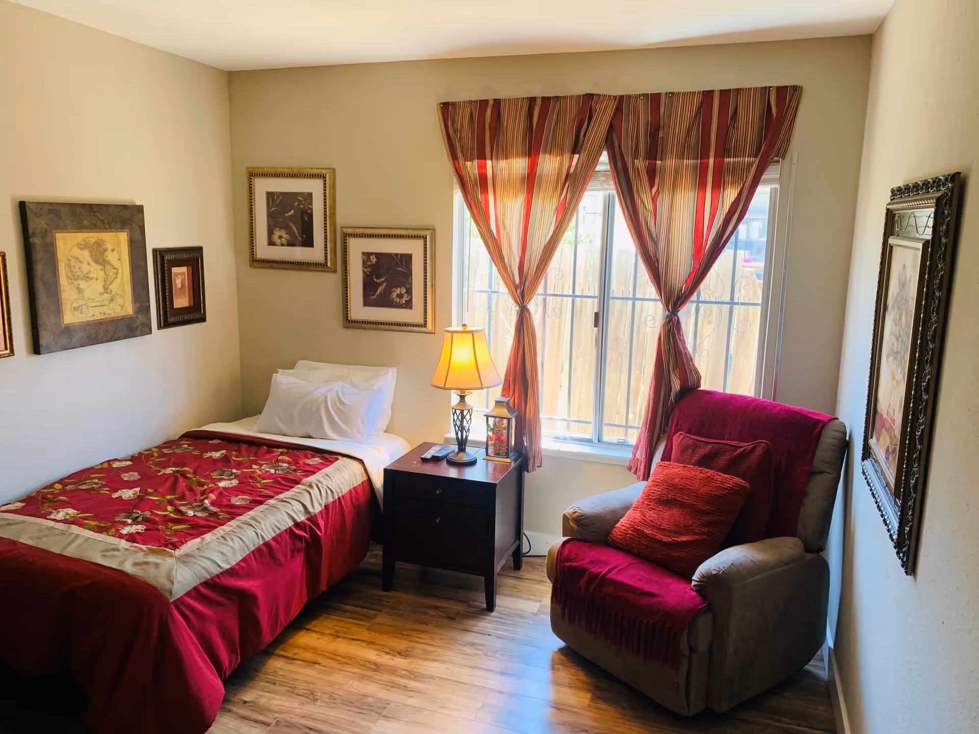 A cozy bedroom with a single bed covered in a red and beige floral bedspread, a nightstand with a lamp and remote controls, a comfortable armchair draped with a red throw and cushion, framed artwork on the walls, and a window with red and beige striped curtains letting in natural light.