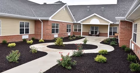 Outdoor courtyard area at Orchard Pointe Health Campus featuring a winding concrete pathway surrounded by landscaped mulch beds with small shrubs and plants, bordered by single-story buildings with beige siding and red brick accents.