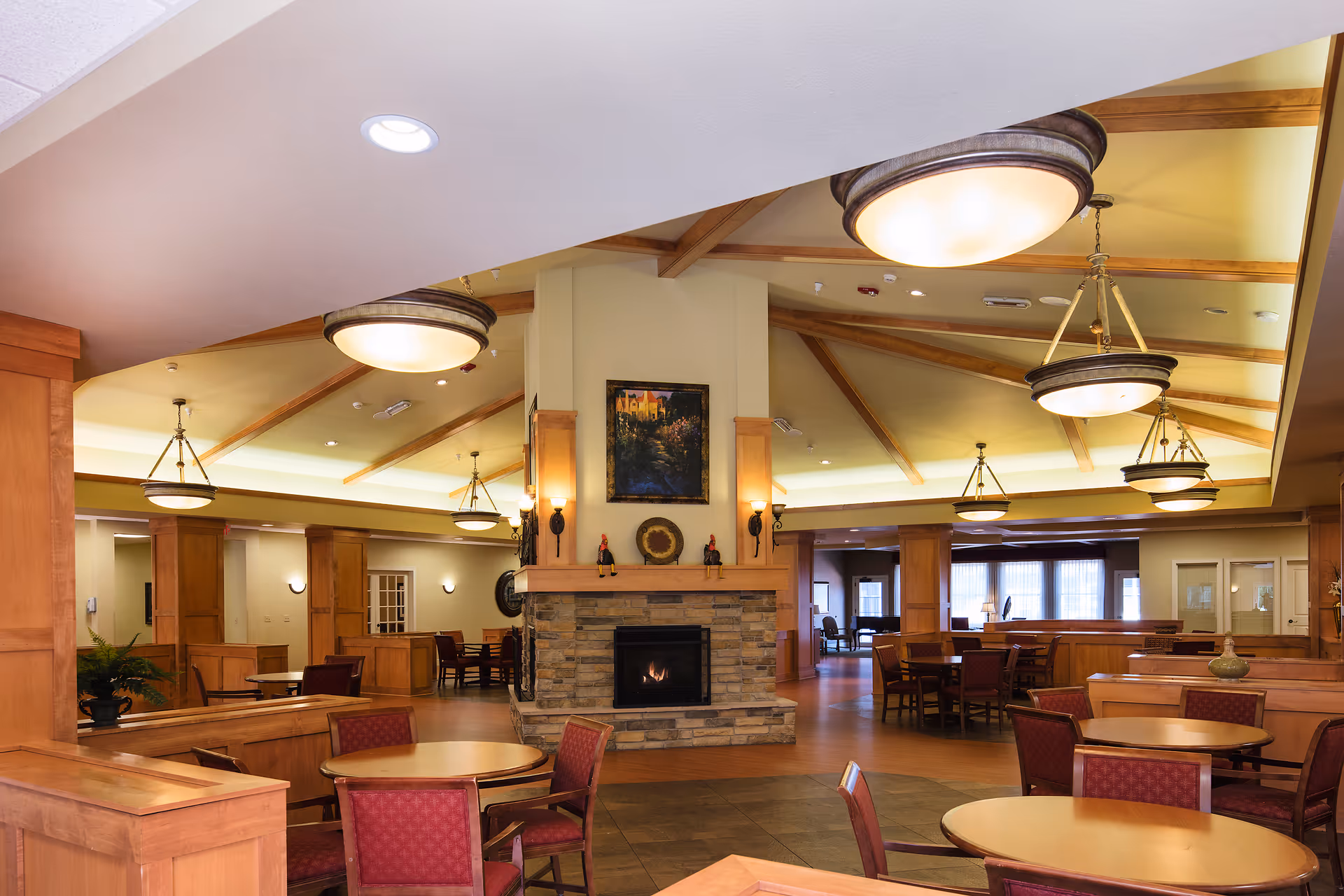 A spacious senior living facility common area with round wooden tables and red cushioned chairs arranged around a central stone fireplace. The ceiling features wooden beams and multiple hanging light fixtures, creating a warm and inviting atmosphere. Large windows in the background allow natural light to fill the room.