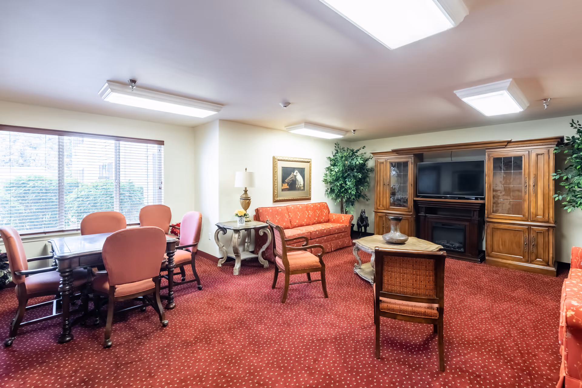 Bright common lounge with red carpet, upholstered chairs and sofas, a TV in a wooden cabinet, and a dining table by the window.
