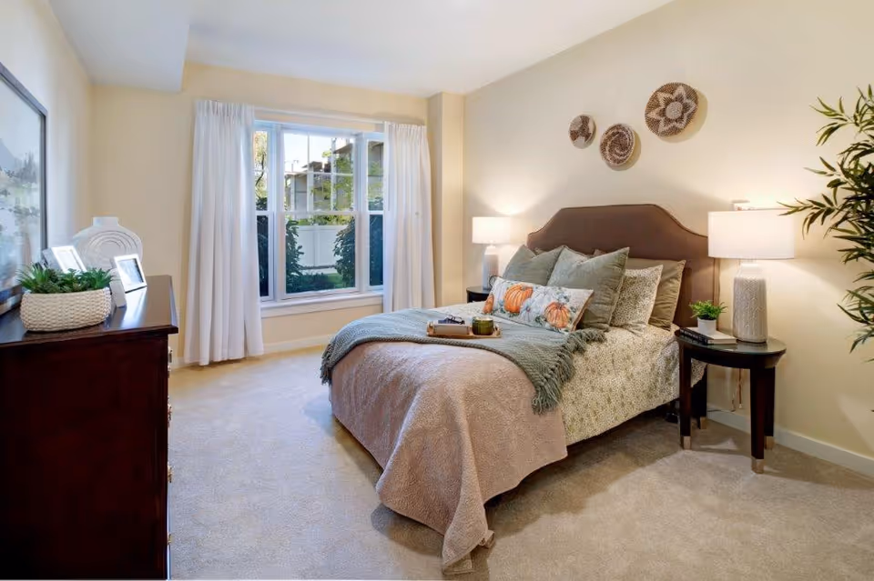 A cozy bedroom with a neatly made bed featuring multiple pillows and a throw blanket. There are two bedside tables with lamps on each side of the bed. A large window with white curtains lets in natural light. A dresser with decorative items and framed pictures is on the left side of the room. The walls are painted a soft beige, and there are three decorative woven wall hangings above the bed.