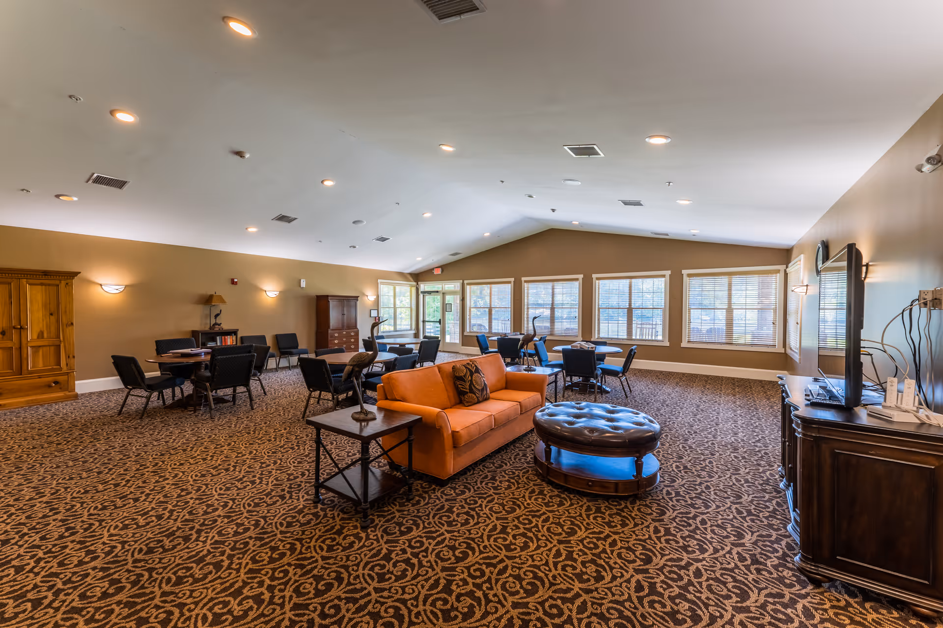 A spacious senior living common area with patterned carpet, an orange sofa, a round tufted ottoman, several tables with chairs, large windows letting in natural light, and a TV on a wooden cabinet.