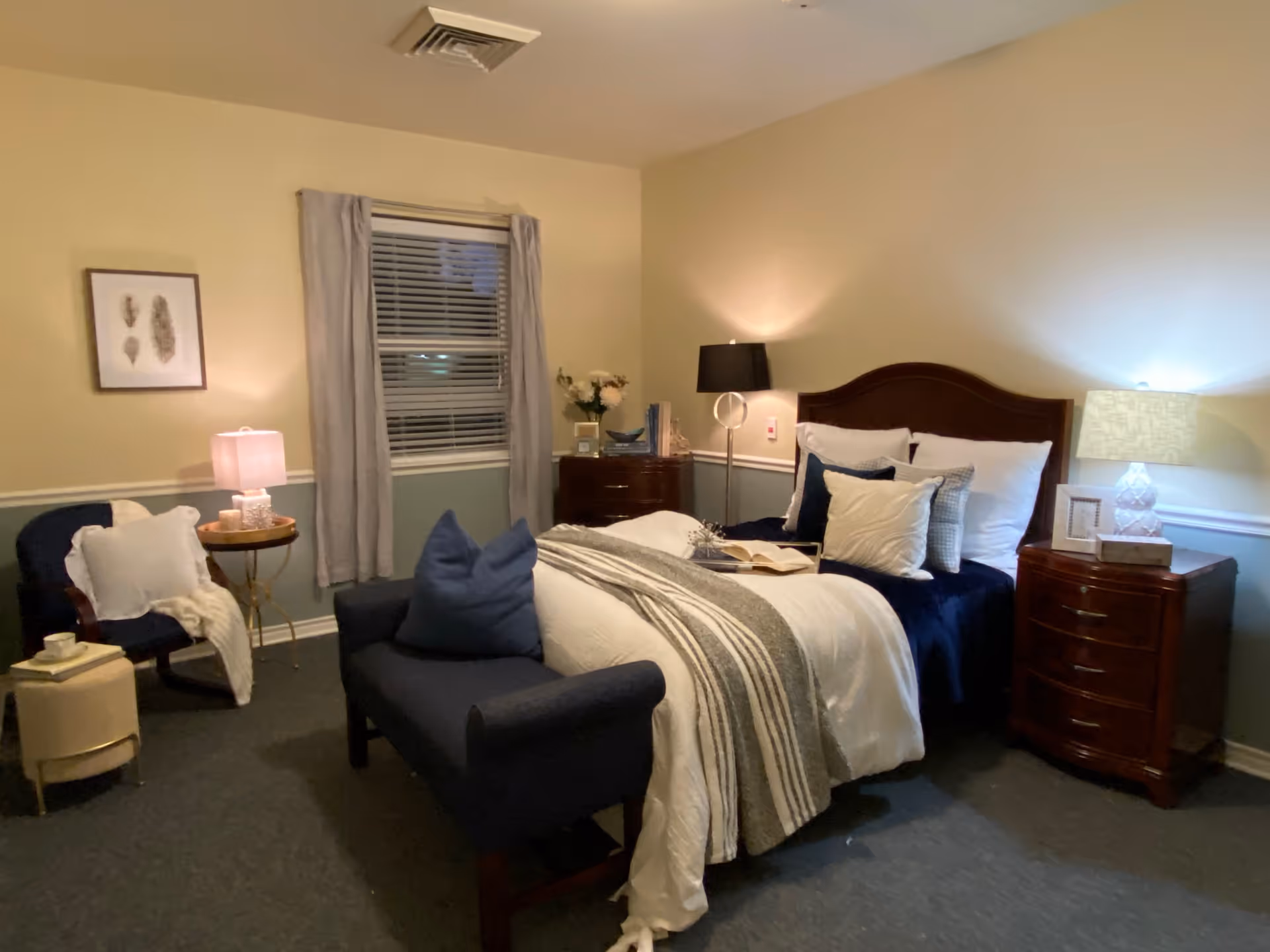 A cozy bedroom with a neatly made bed featuring white and navy blue bedding and multiple pillows. There is a dark wooden headboard and two matching nightstands with lamps on either side of the bed. A small navy blue loveseat is placed at the foot of the bed. To the left, there is a blue armchair with a white throw blanket and a small round side table with a lamp. A window with closed blinds and gray curtains is centered on the back wall. The walls are painted beige with a white chair rail and a muted green lower section. The room is softly lit with warm lighting.