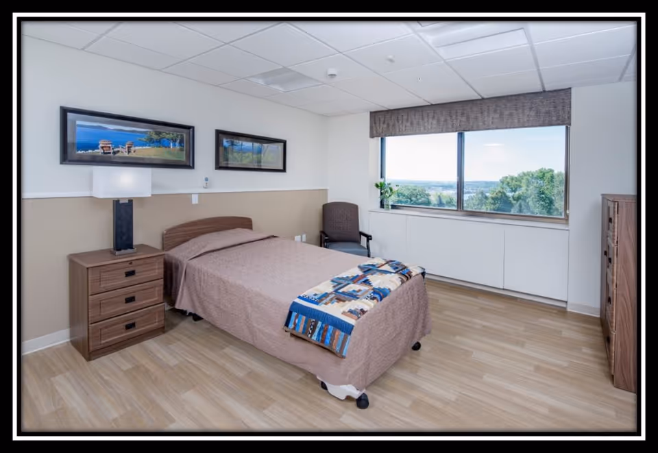 A clean and well-lit bedroom in a senior living facility featuring a single bed with a brown bedspread and a colorful quilt at the foot. Next to the bed is a wooden nightstand with a modern lamp. The room has light wood flooring, a large window with a view of trees and a distant landscape, a comfortable chair, and two framed landscape pictures on the wall.