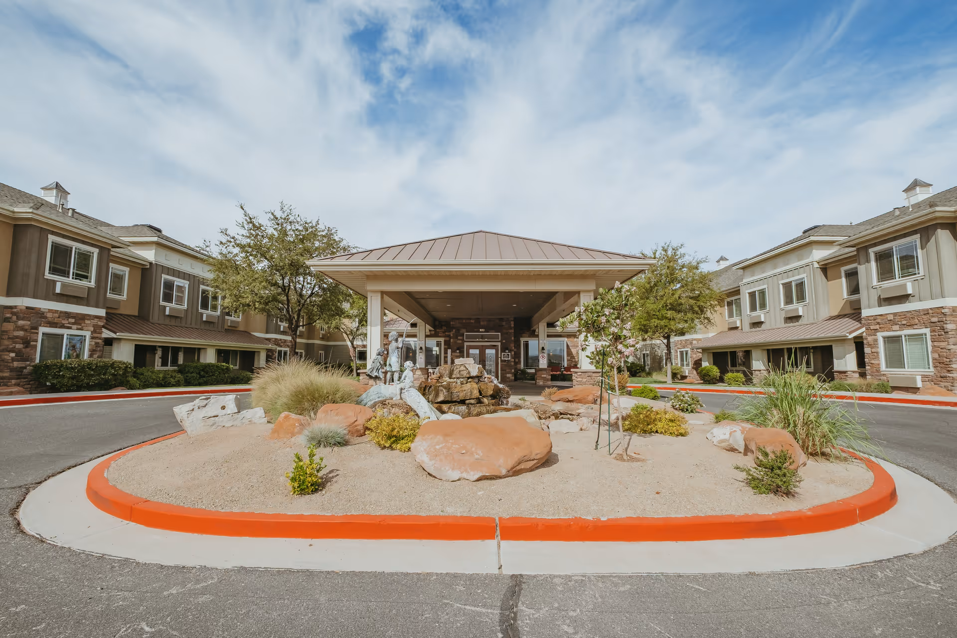 Front entrance of Spring Gardens St. George facility with a covered drop-off area, landscaped roundabout featuring rocks, plants, and a statue of children playing near a water fountain, flanked by two-story buildings under a partly cloudy sky.