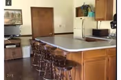 Interior view of a kitchen area with a long countertop island surrounded by six wooden bar stools. The kitchen features wooden cabinets, a white refrigerator, a microwave, and a sink. There is a wooden door and a small TV on a stand in the background.