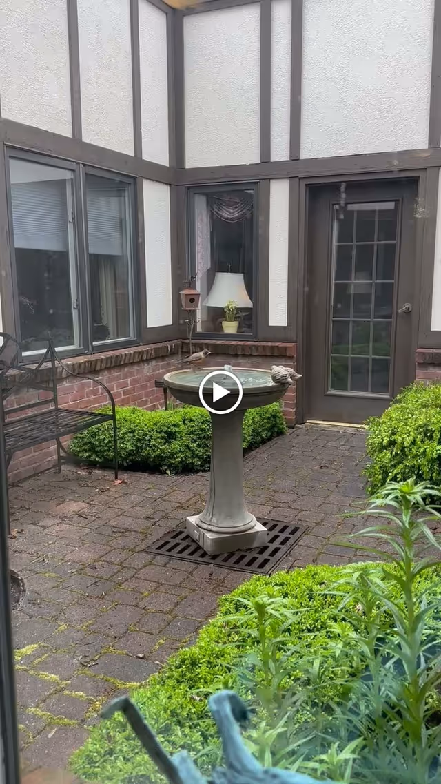 Enclosed brick courtyard featuring a pedestal birdbath fountain, metal bench, trimmed shrubs, and adjacent windows and a door.