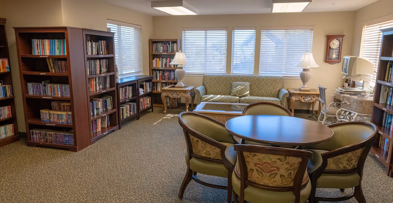 A cozy senior living community room with multiple bookshelves filled with books, a round wooden table surrounded by four upholstered chairs, a green patterned sofa, two side tables with lamps, a wall clock, and a computer desk with a chair near windows with blinds.