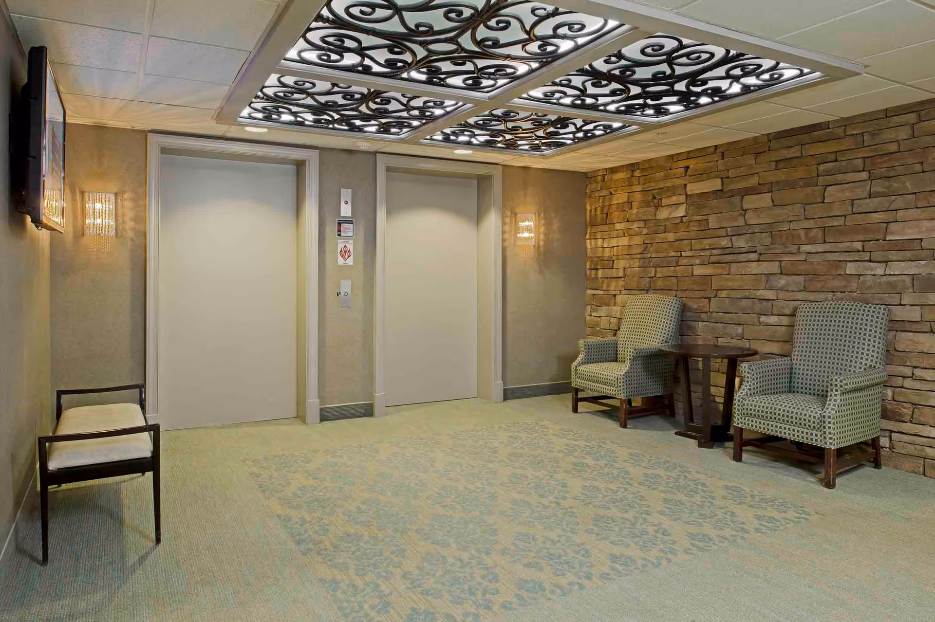 Interior view of a hallway area with two closed elevator doors, a stone accent wall, two patterned armchairs with a small wooden table between them, a bench on the opposite side, and decorative ceiling panels with intricate black scrollwork.