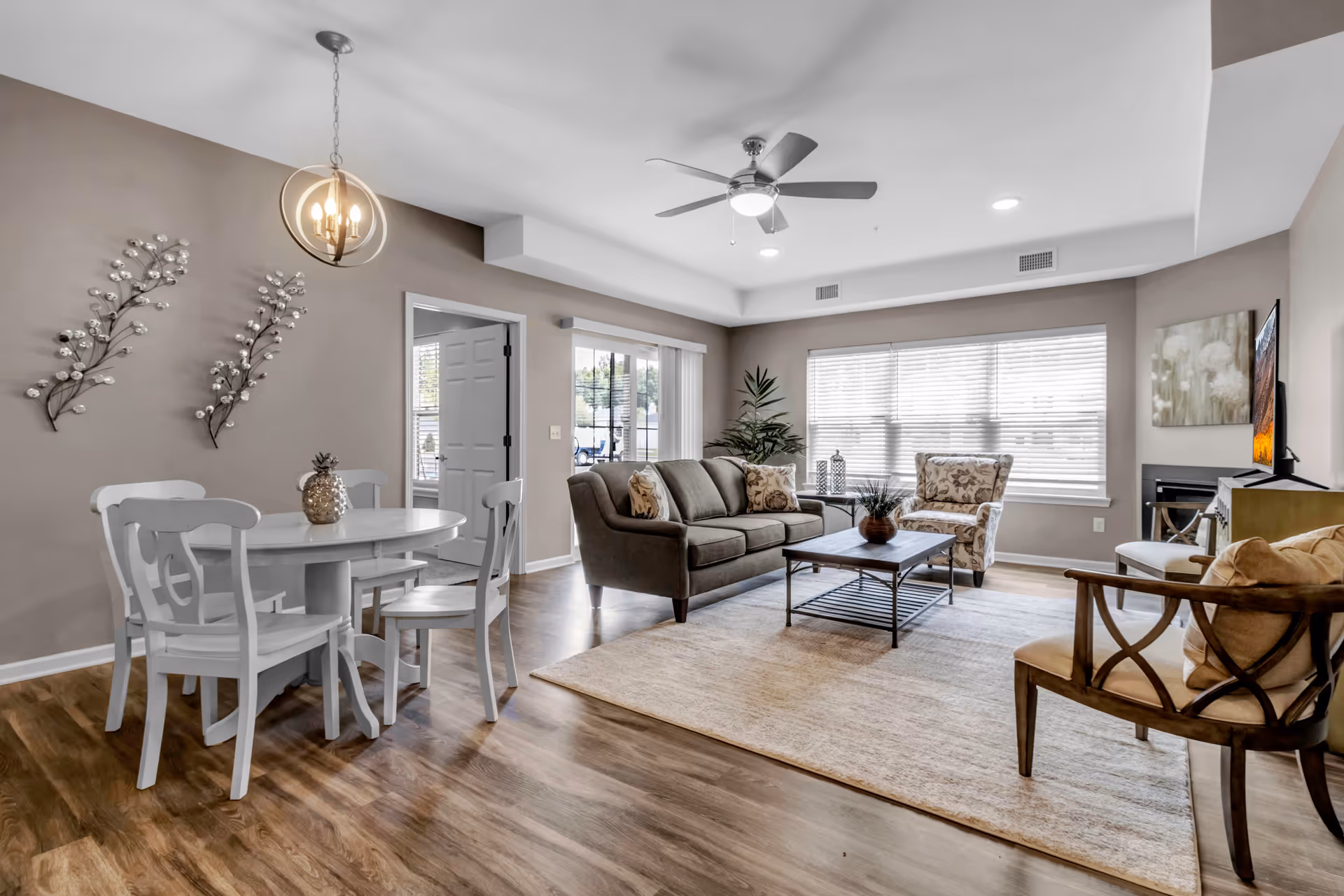 A bright and spacious living room and dining area with wooden flooring. The living room features a gray sofa with patterned cushions, a floral armchair, a wooden coffee table, and a TV mounted on the wall above a fireplace. The dining area has a round white table with four matching chairs and a decorative pineapple centerpiece. The walls are painted beige with two metal wall art pieces above the dining table. Large windows with blinds allow natural light to fill the room, and a ceiling fan with a light fixture is mounted on the ceiling.