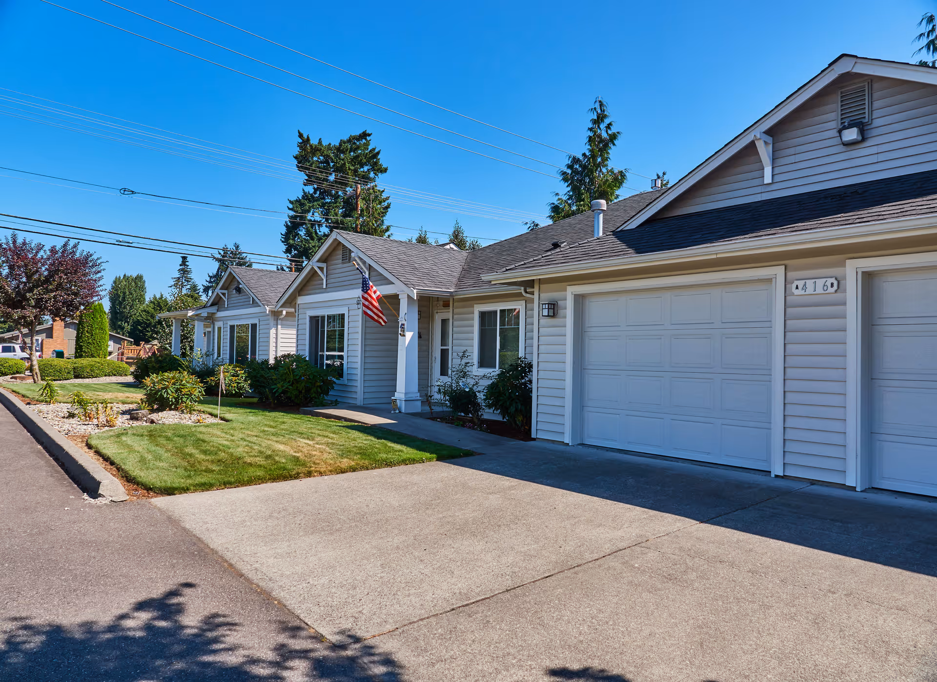 Exterior view of a single-story residential building with light gray siding, white trim, and a garage. There is a small front lawn with bushes and an American flag mounted near the entrance. The sky is clear and blue.