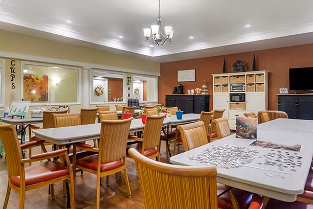 A well-lit activity room with several tables and wooden chairs with red cushions. One table has a partially completed jigsaw puzzle and puzzle pieces scattered on it. The walls are painted in warm tones with decorative items and storage cabinets along the back wall. There is a chandelier hanging from the ceiling and windows looking into adjacent rooms.