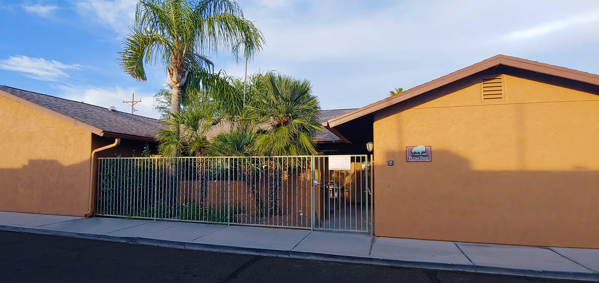 Single-story stucco building front with a gated entrance, palm trees, and a sidewalk under a clear sky.