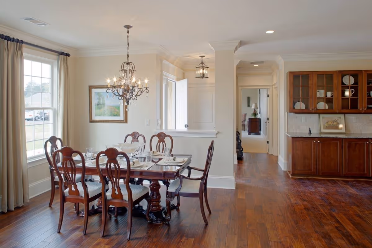 A dining room with a wooden dining table set for six, featuring elegant chairs with carved backs. A decorative chandelier hangs above the table. The room has large windows with beige curtains allowing natural light to enter. To the right, there is a built-in wooden cabinet with glass doors displaying dishes and decorative items. The floor is made of polished wood, and the walls are painted a light cream color.